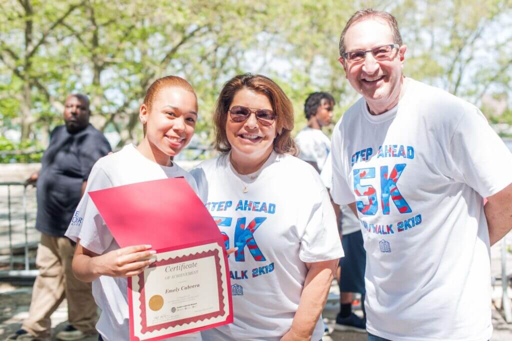 David and Andrea Acker pose for a photo with a CBI student who is holding a certificate for completing the Step Ahead 5K in 2019.