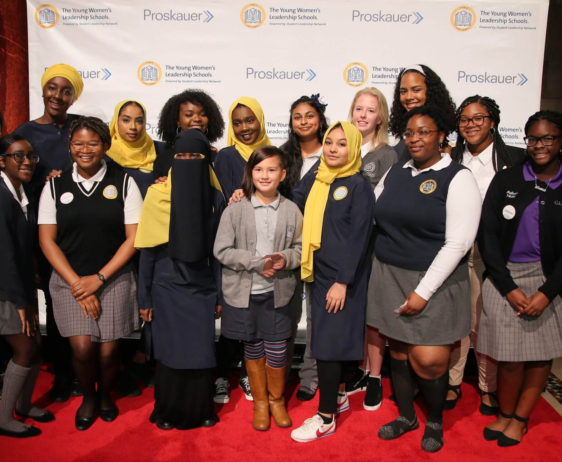 Group of smiling girls wearing a variety of school uniforms stand in front of a step and repeat with logos for The Young Women's Leadership Schools and Proskauer