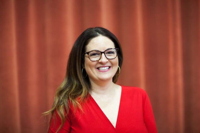 Jessica Kane is standing in front of a red curtain. She is smiling.