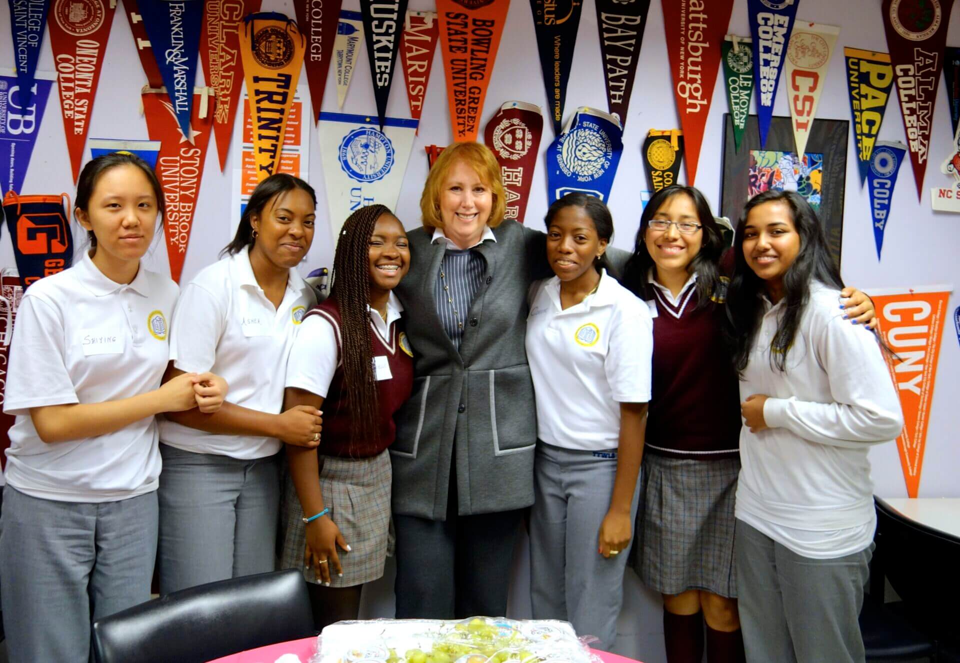 Ann Tisch is standing with students in their college office. Colorful college pennants line the wall behind them.