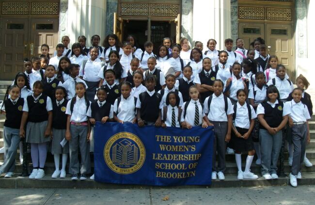 The first class at TYWLS Brooklyn stands on the steps to the school. The front row is holding a blue banner that reads "The Young Women's Leadership School of Brooklyn."