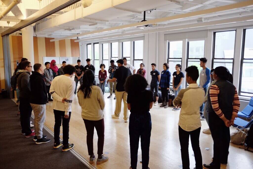 A male volunteer stands in the middle of circle of students, young men and women, also standing