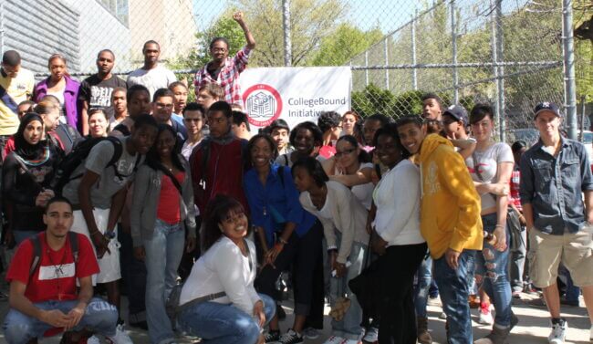 A group of about 30 CBI students stand in front of a fence and CBI sign.