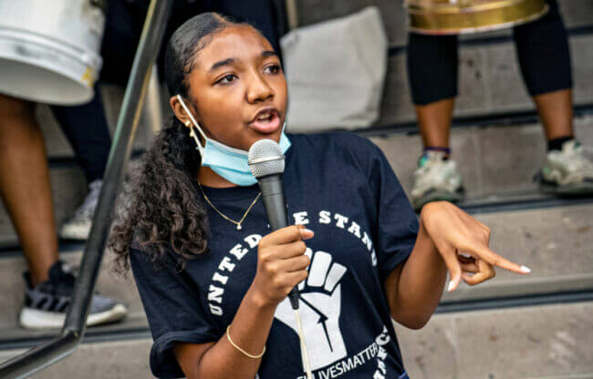 Kahlila is standing on steps and holding a microphone, speaking at a Black Lives Matter event.