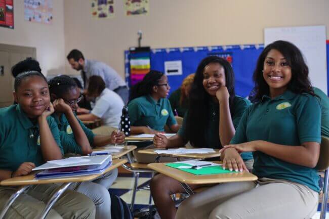 Four students from YWCP are sitting in a classroom with their desks facing each other. They are taking a break from their group homework to smile for the camera.
