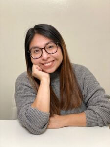 Alendi is leaning on a desk with her chin propped on her hand. She is smiling warmly at the camera. 