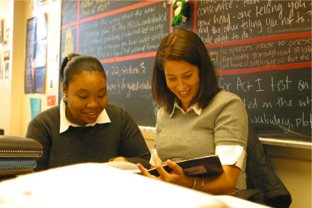 Lindsay Gruber Dunham back in the day when she taught at TYWLS East Harlem. Here she is sitting with a student looking at a book. They are both smiling.