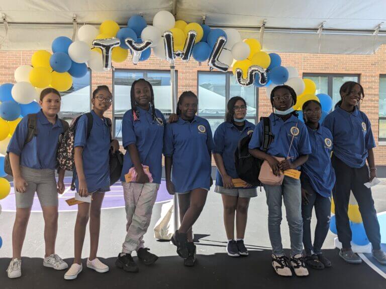 A group of eight students stand beneath an arch made of blue and yellow balloons. TYWLS is spelled out in silver balloons.