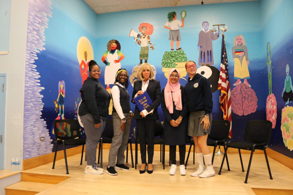 First Lady Macron stands in the middle of the four TYWLS students who moderated the panel discussion. Macron is holding a copy of "Its all about the girls"
