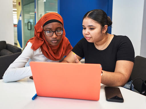 Two young women, both CollegeJourney peer leaders, sit at a table focused on a coral-colored laptop. One wears a red hijab and glasses, while the other wears a black top. A smartphone rests nearby on the table.