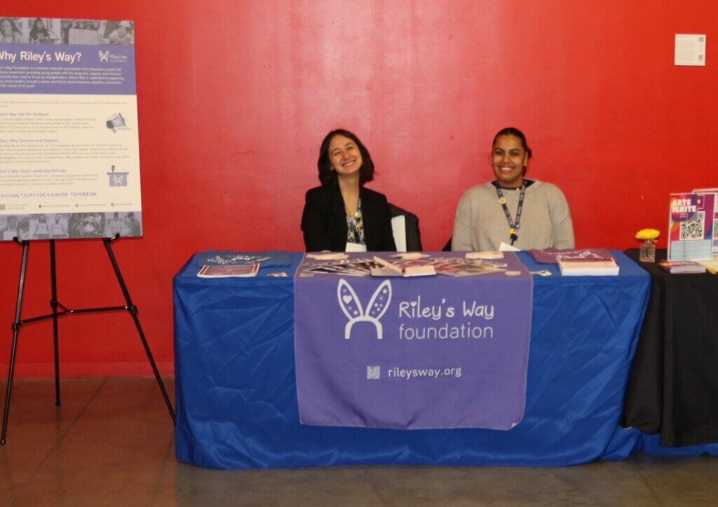 Two smiling young women sit behind a Riley's Way exhibitor table