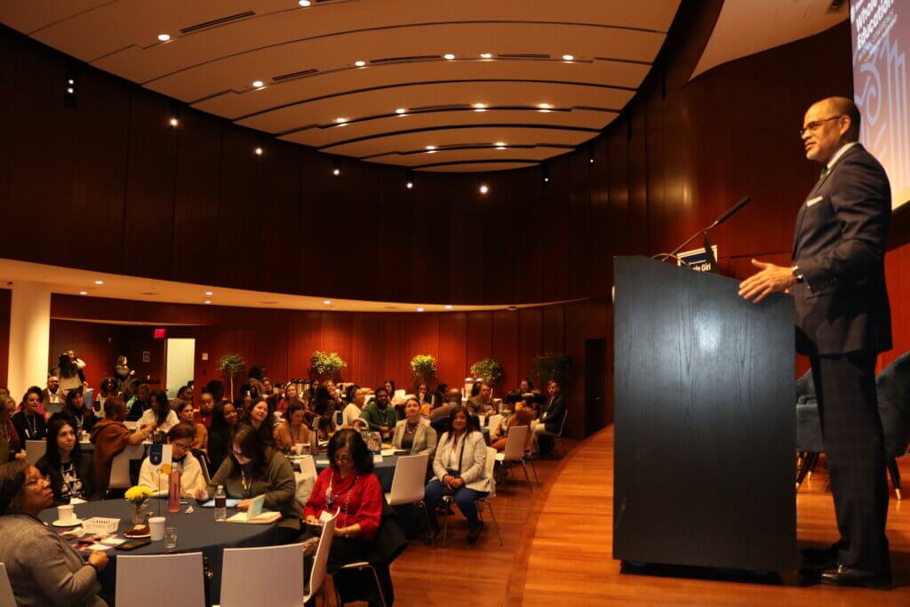 David Banks stands behind a podium on stage addressing a roomful of conference attendees