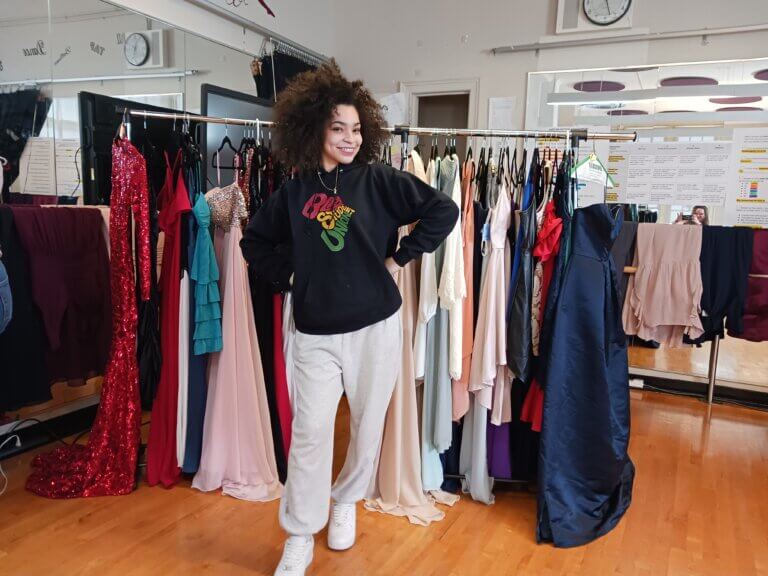Alumna Symphony J. stands in front of a rack of donated prom dresses