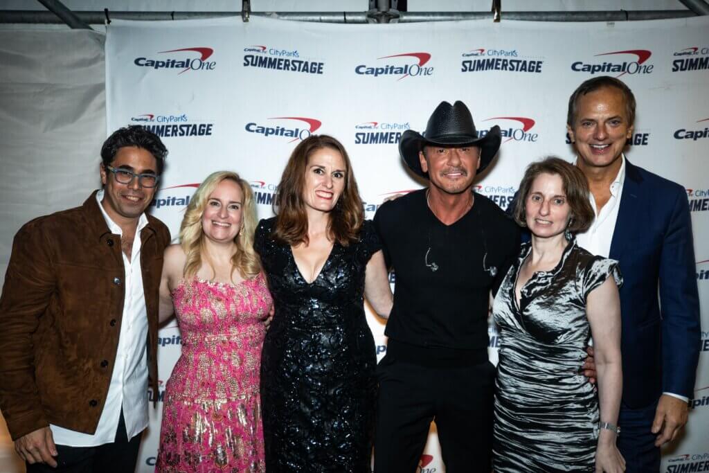 Six adults in formal wear pose for photo, with backdrop of logos for "City Parks Summer Stage"