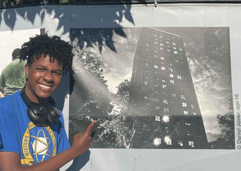 Student Feature: In front of a wall of public artwork, a young man points to a black and white image of a tall building he had photographed