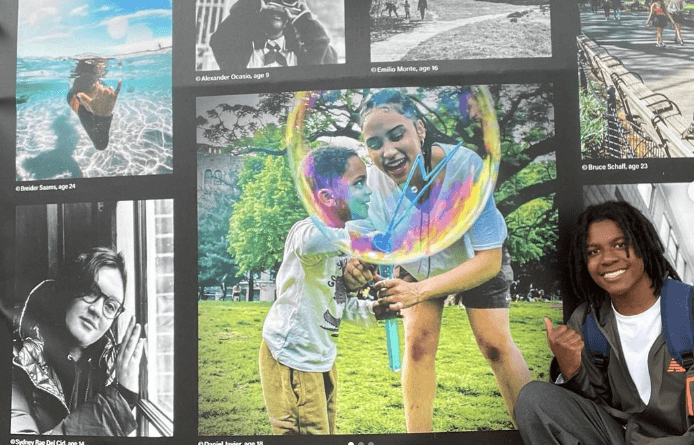 In front of a wall of public artwork, a young man points to an image of a caregiver and child that he had photographed