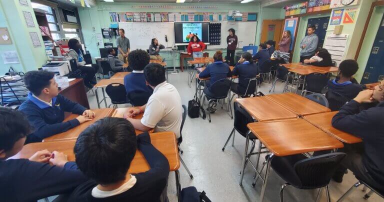 Students in navy blue uniforms are seated, facing front of classroom, where teacher and alumni are presenting