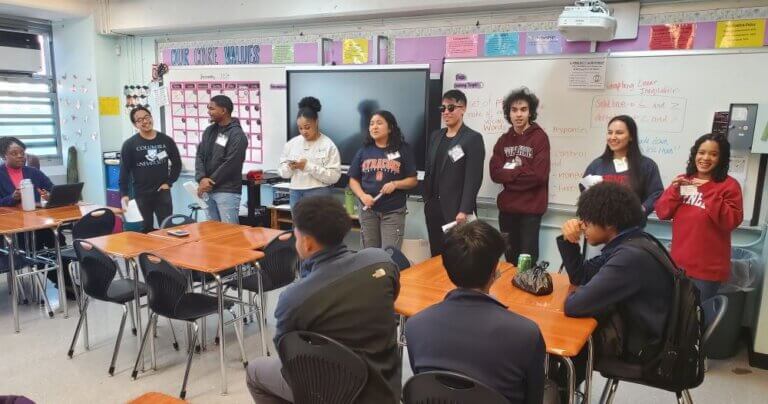 Eight alumni wearing college sweatshirts stand at front of classroom to speak with seated students