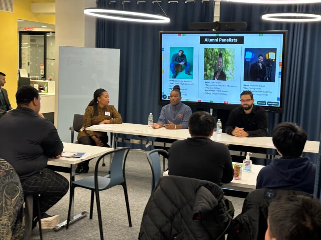Three adults have a panel discussion at front of the room, with students seated facing them
