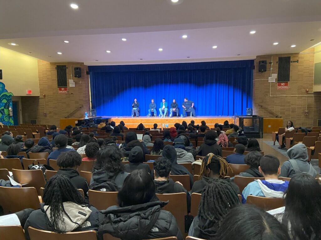 Alumni Days: Alumni and college counselor are seated on stage, with a filled auditorium of seated students