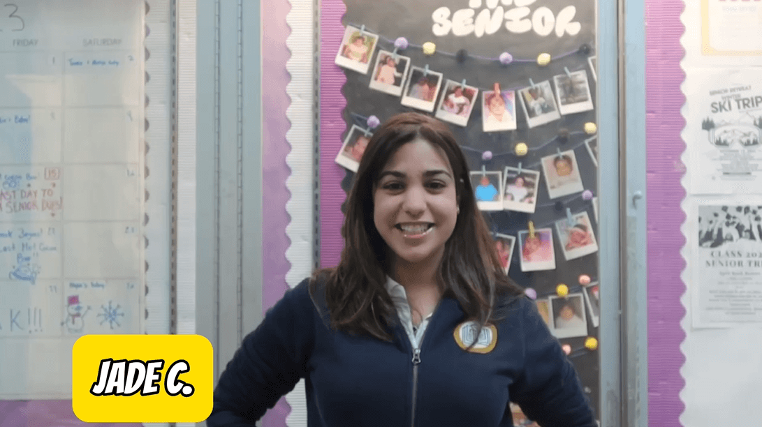 Student wearing school uniform sweater stands in front of a bulletin board of senior photos