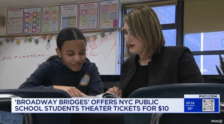 Student and teacher look at a book together in classroom, with news banner on bottom of screen