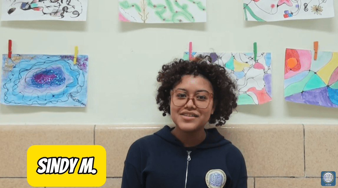 young woman with curly hair and glasses stands in front of artwork wall
