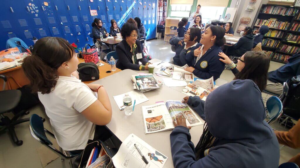 Students and adult volunteer sit at classroom tables, looking over a stack of magazines the volunteer brought.