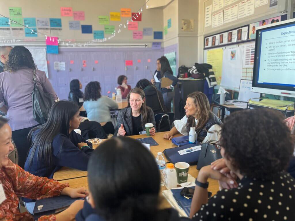 Students and adult volunteers talk with each other while seated around school desks