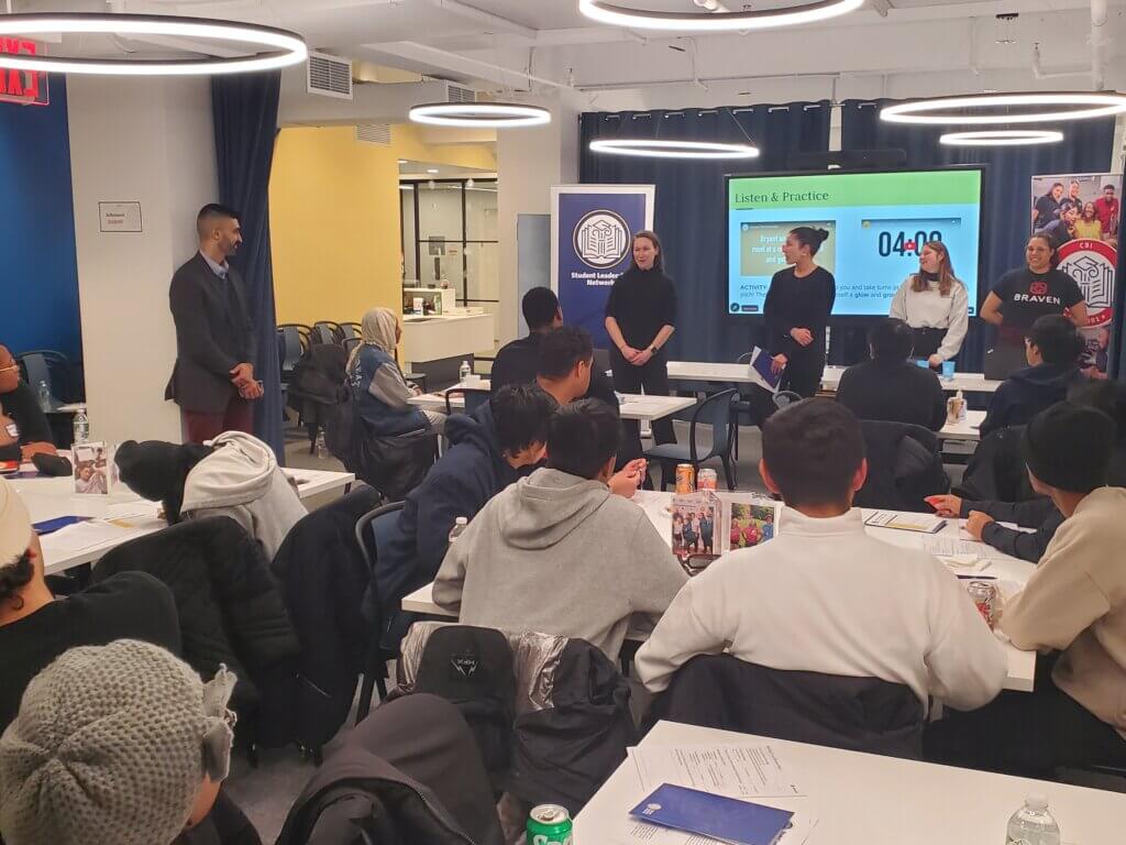 Four adults present in front of room while students and alumni are seated at small tables