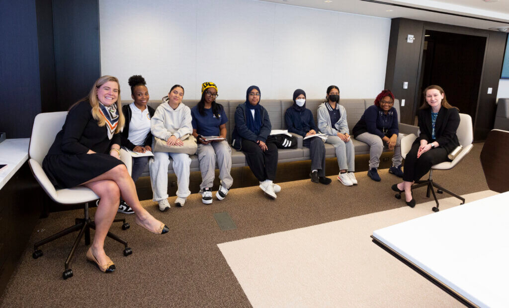 Students are seated on a long couch inside an office, with two adult volunteers seated in chairs on opposite ends