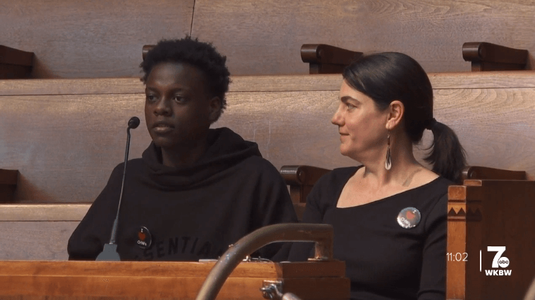Young Black male student speaks into a podium microphone, with a school administrator seated next to him