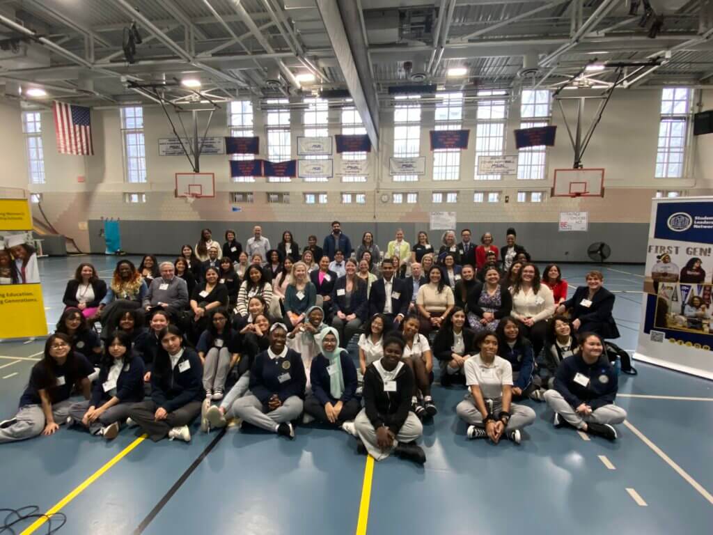 Large group of students and adult volunteers are seated for photo in gymnasium