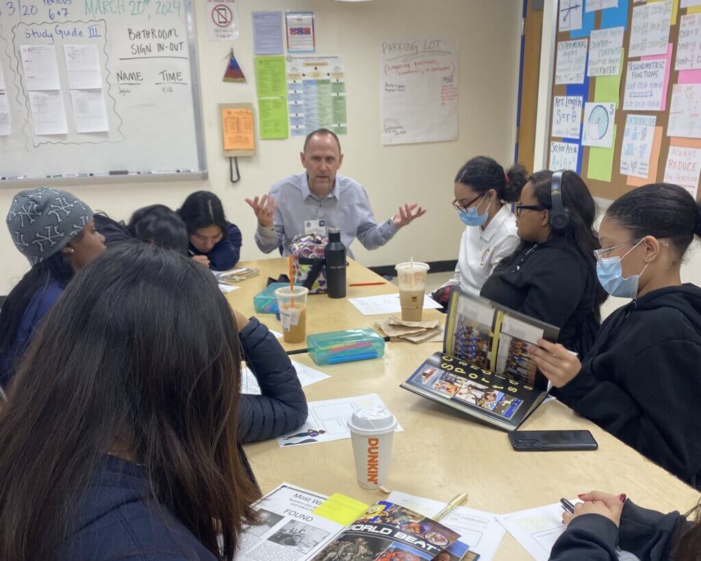Adult volunteer gestures with hands as he speaks with a group of students around a table. Students are looking at yearbooks while he speaks.