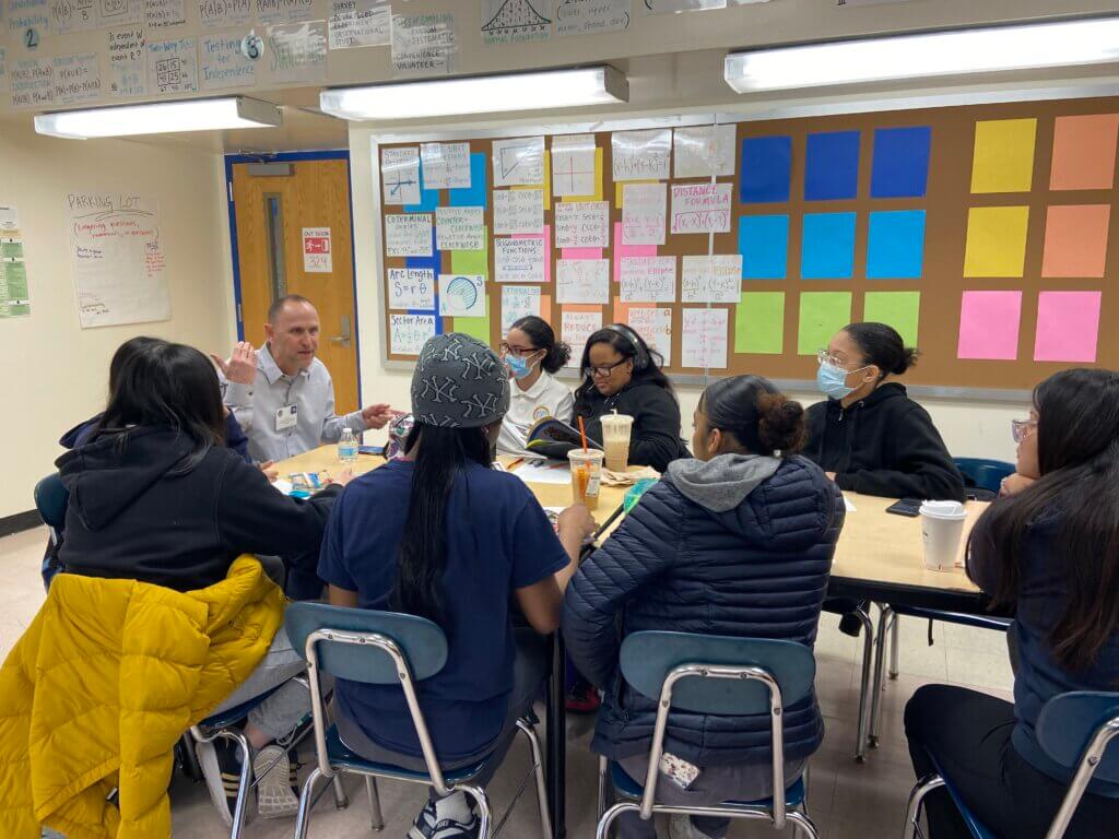 Adult volunteer sits at head of oval shaped table with students