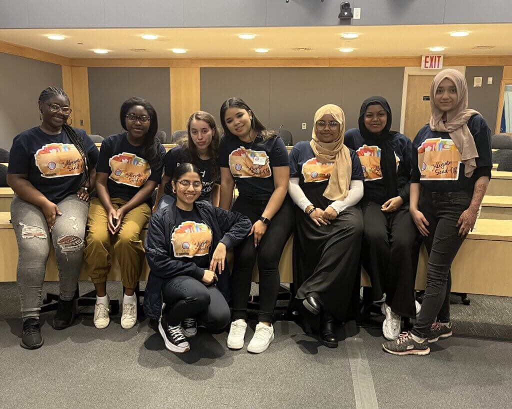 Eight young women in matching "Collegiate Send Off" t-shirts pose for a photo in an auditorium