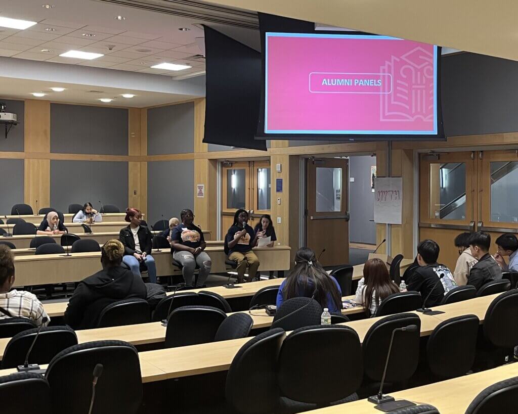 Group of alumni seated at front of room talk to students in an auditorium, with projected screen with text "Alumni Panels"