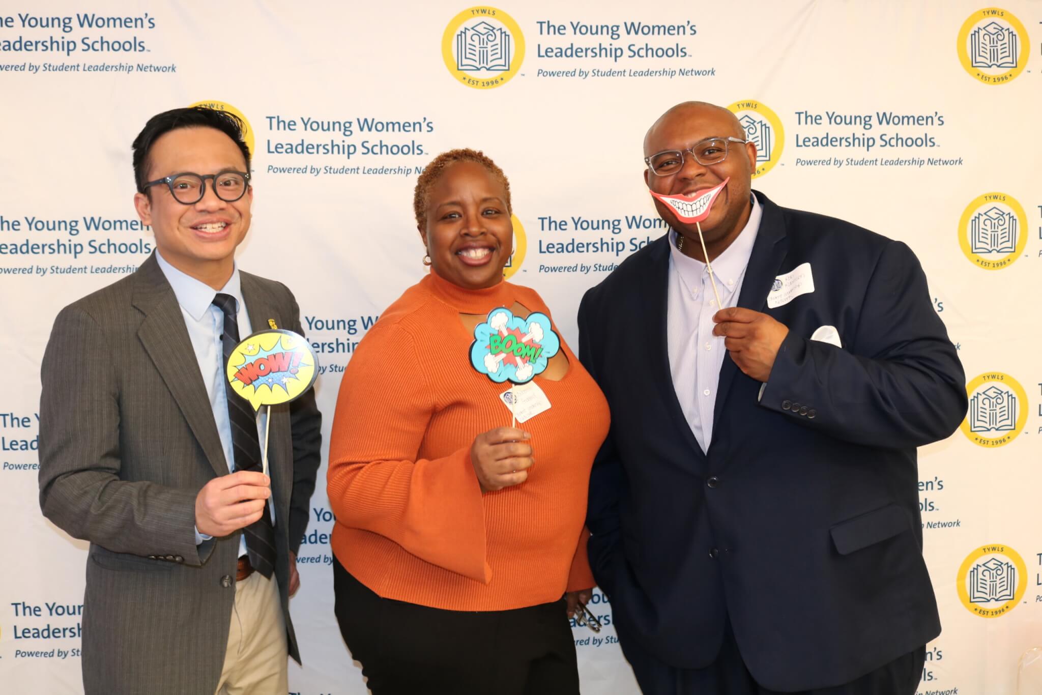 Three adults hold up colorful graphic cutouts on sticks, posing for photo in front of a company logos backdrop