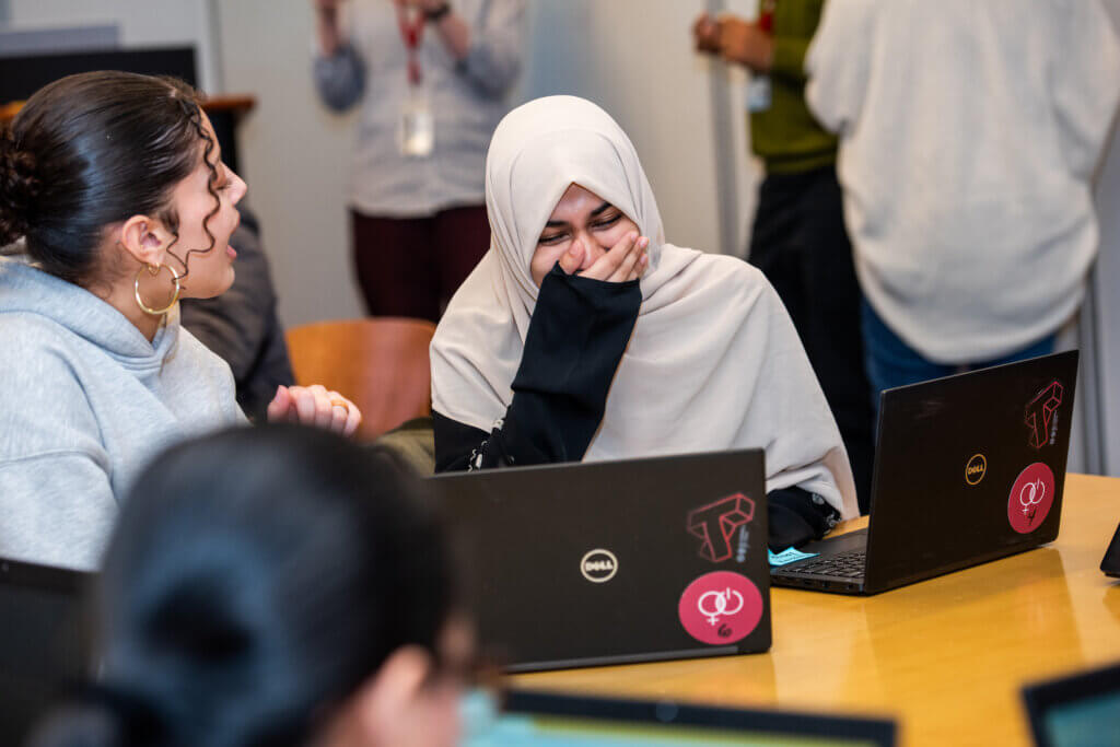 Student holds hand to her mouth as she laughs, while another student speaks to her on the side, both have laptops open