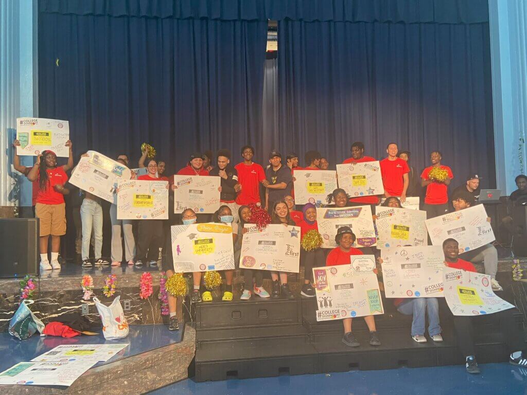High school seniors hold up college posters on stage