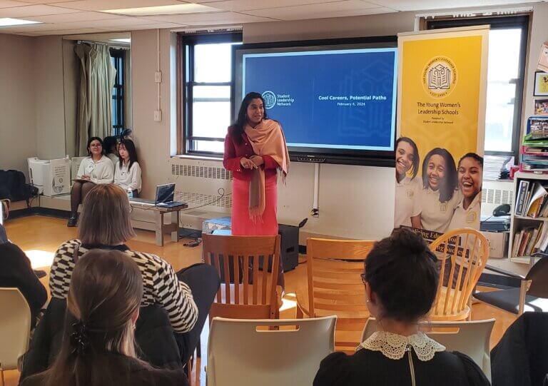 Principal stands in front of room to welcome volunteers, adults are seated in front of her and students on the side