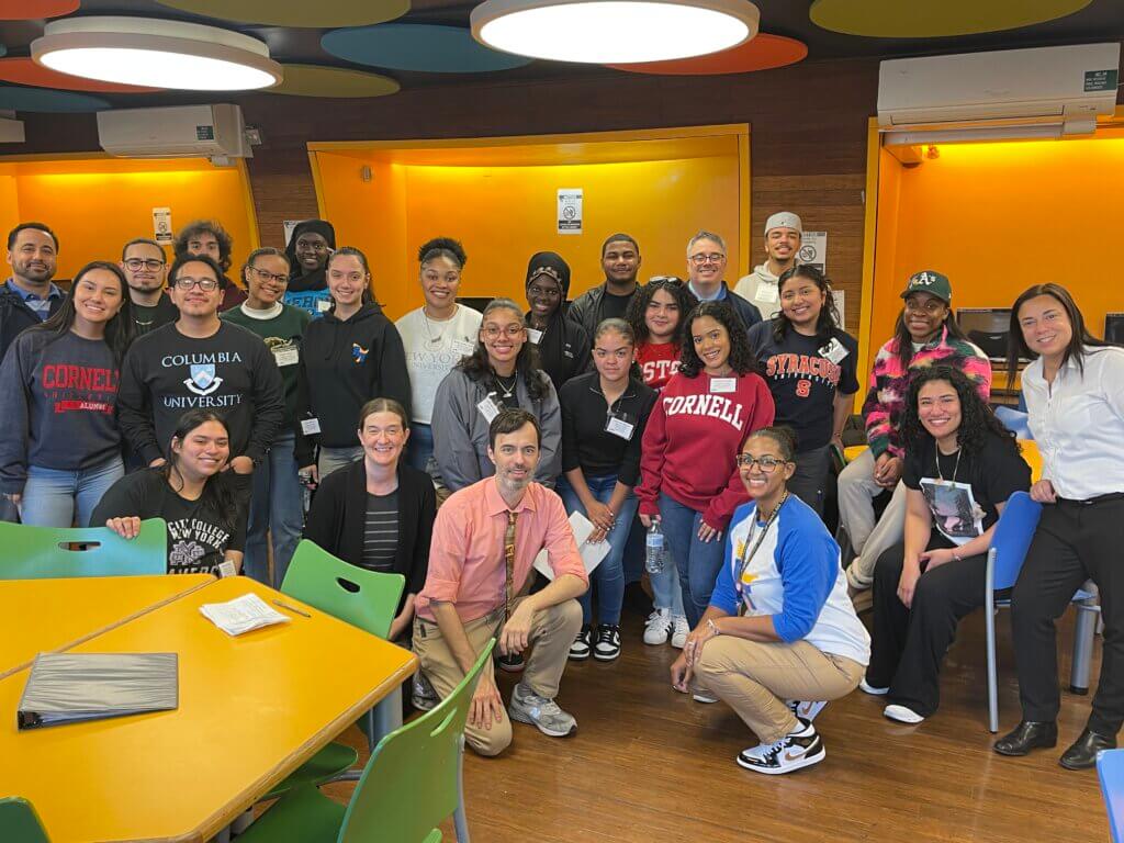 Alumni wearing sweatshirts with the names of their colleges pose for a photo with teachers