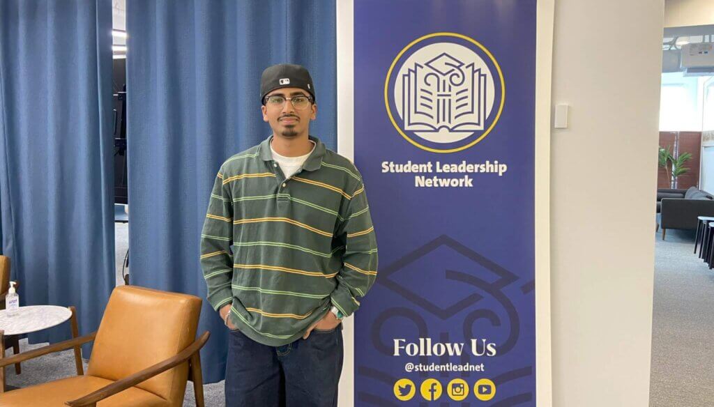 Young man wearing a cap, glasses, and a striped shirt poses for photo next to Student Leadership Network sign
