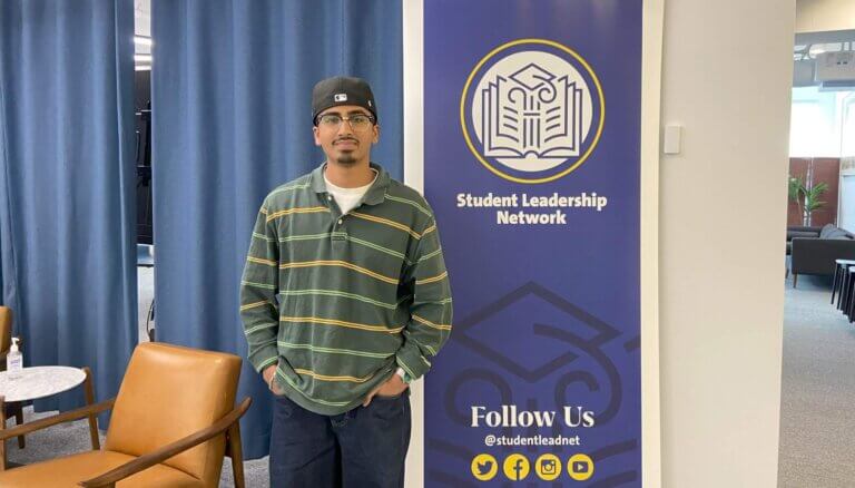 Young man wearing a cap, glasses, and a striped shirt poses for photo next to Student Leadership Network sign