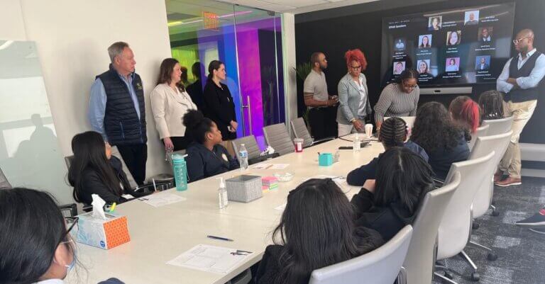 Adult volunteers gather at front of conference room by slideshow tv and students are seated at table
