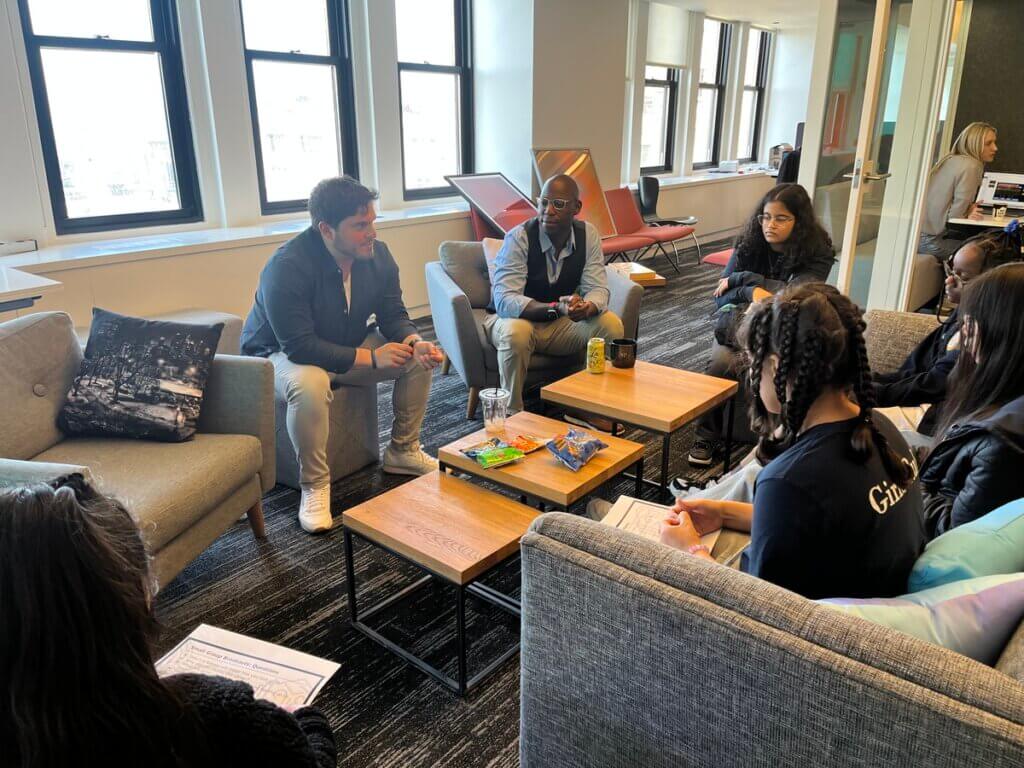 Two adults and four students sit on individual couches in an office space