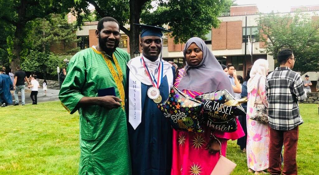 Young Black man in a graduation cap and gown, wearing two medals, stands with his parents in their cultural dress on a lawn
