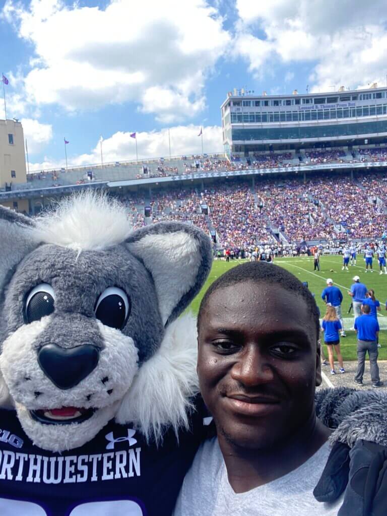 Young man poses with a football mascot a wolf with a football stadium in the background