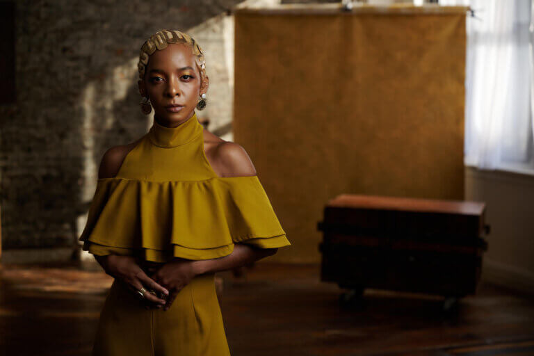 A young woman Kara Young looks to the front with a confident gaze, wearing a yellow dress and with her hands clasped in front of her