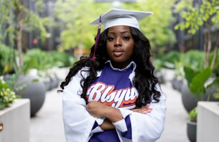 Young woman wears a white graduation cap and gown, with purple t-shirt with text "BLSYW" across the front